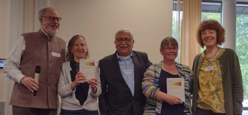 A group of five people standing indoors. Two individuals are holding copies of a book. The person to the far left is holding a microphone.