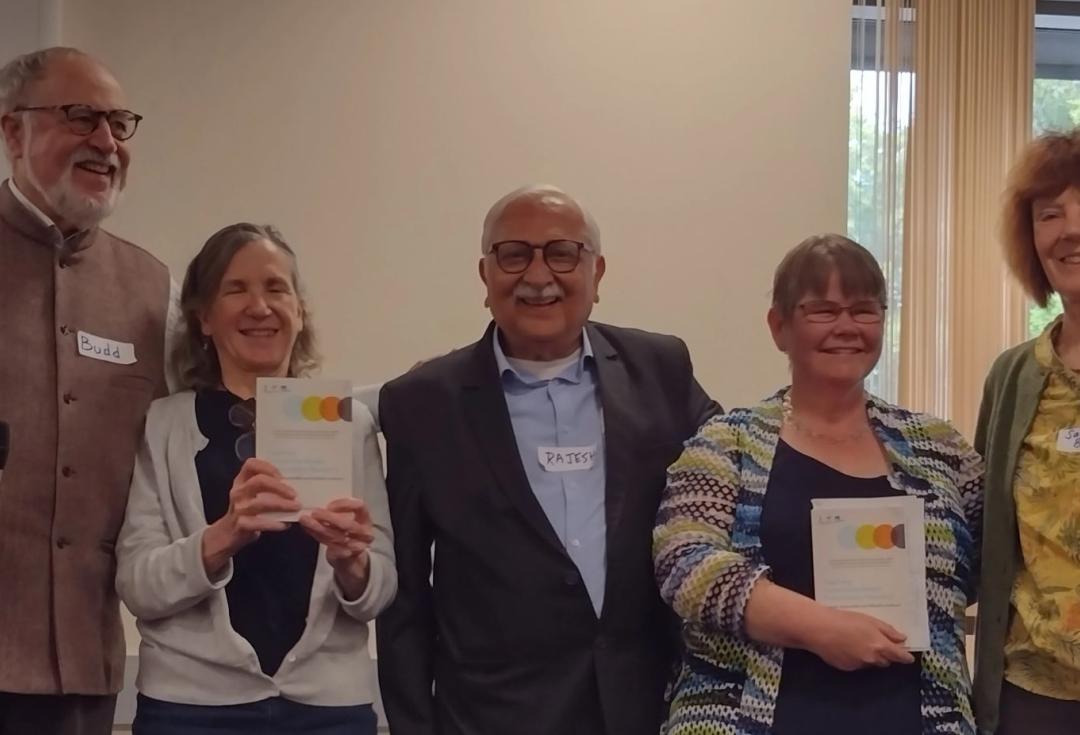 A group of five people standing indoors. Two individuals are holding copies of a book. The person to the far left is holding a microphone.