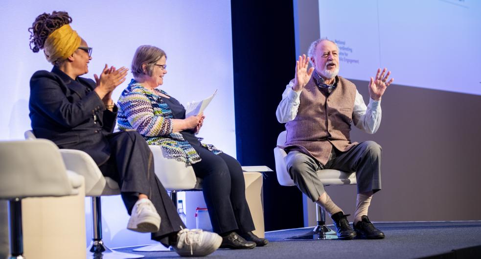 Budd Hall sitting, talking animatedly on stage at Engage with Sophie Duncan and Ima Jackson 