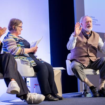 Budd Hall sitting, talking animatedly on stage at Engage with Sophie Duncan and Ima Jackson 