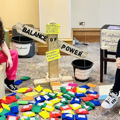 Emily and her colleague sitting on stools either side of Emily's 'balance of power' wooden scales, surrounded by colourful beanbags with labels attached.