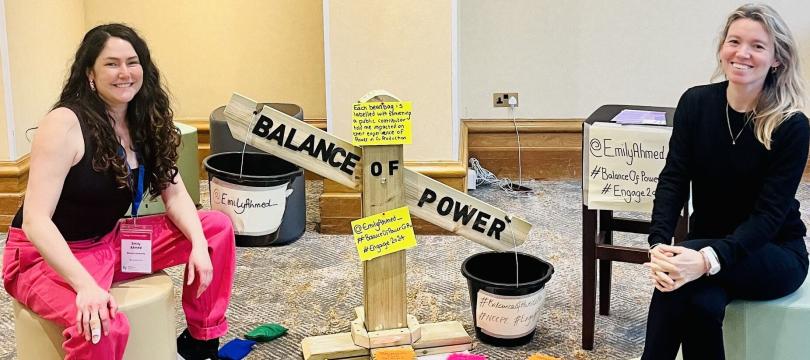 Emily and her colleague sitting on stools either side of Emily's 'balance of power' wooden scales, surrounded by colourful beanbags with labels attached.