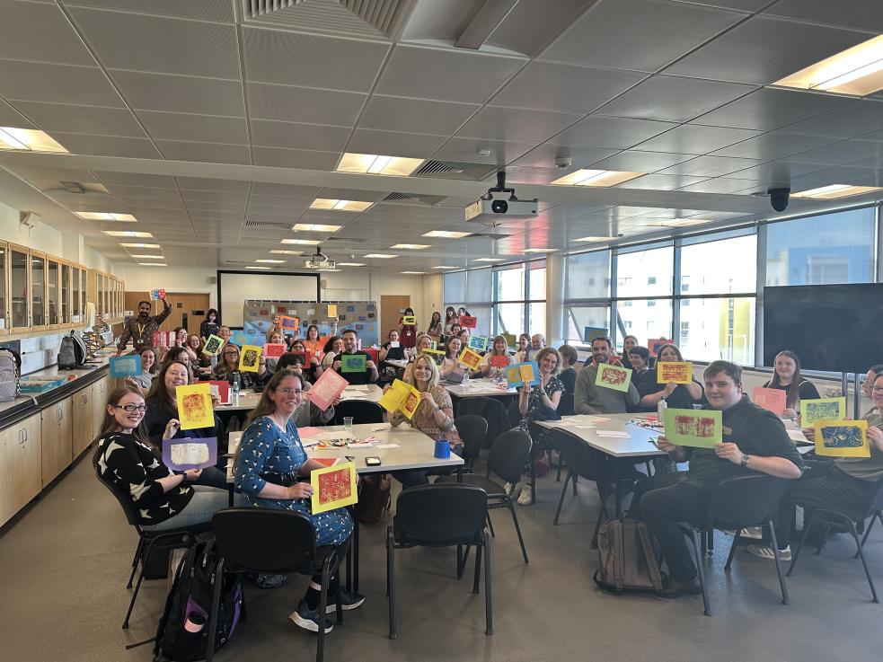 A classroom of people sat at tables holding up printed artwork
