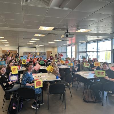 A classroom of people sat at tables holding up printed artwork