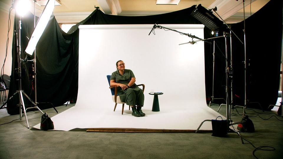 Researcher in a recording studio sat in a chair in front a screen with microphones ready to be filmed
